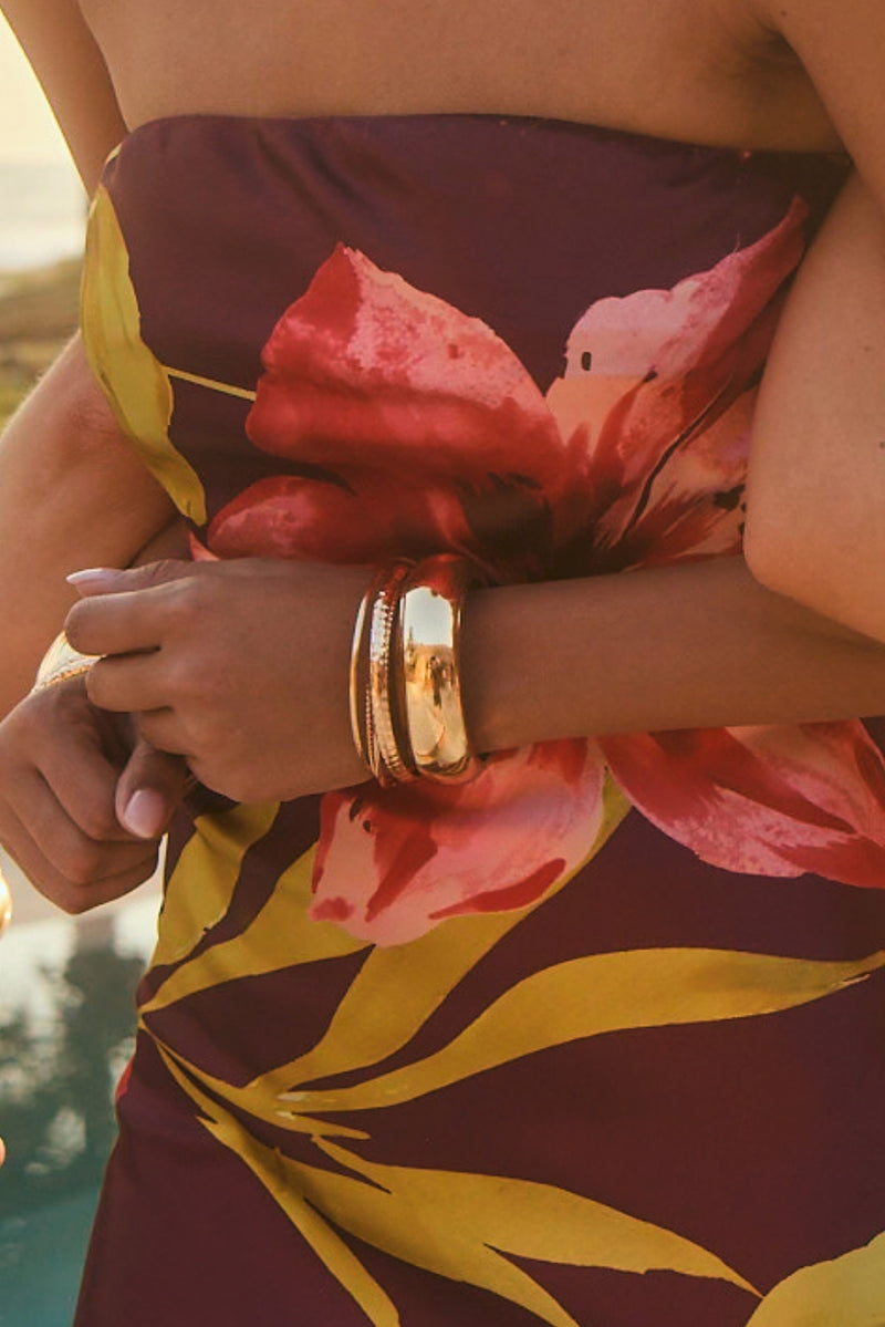 Stacked gold bangle bracelets on a wrist against a floral strapless resort dress.