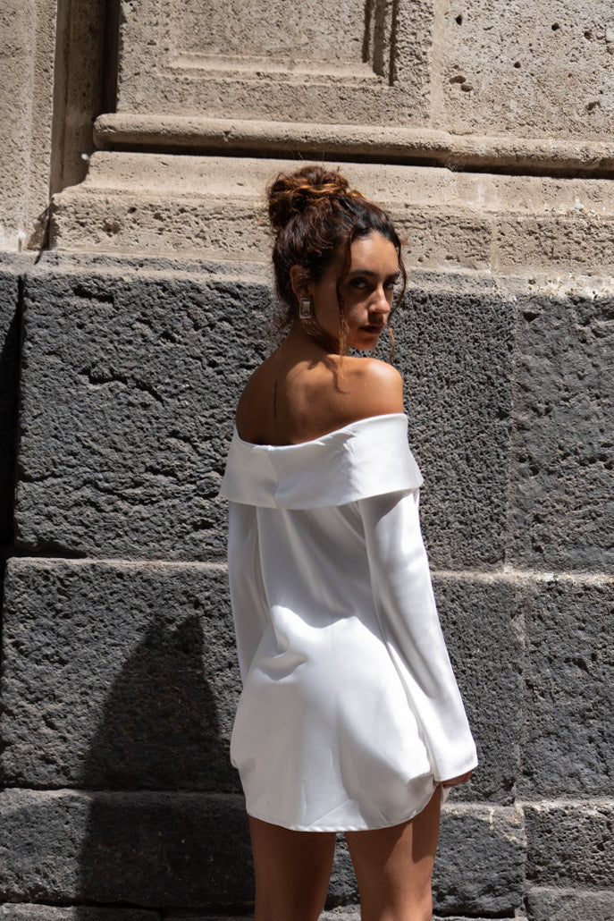 White off-shoulder satin mini dress shown from behind against a stone wall.