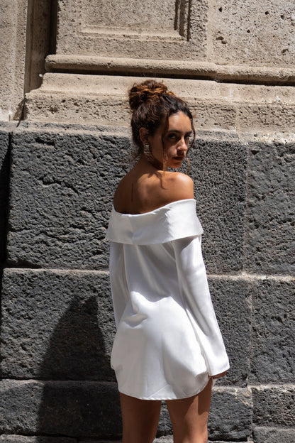 White off-shoulder satin mini dress shown from behind against a stone wall.