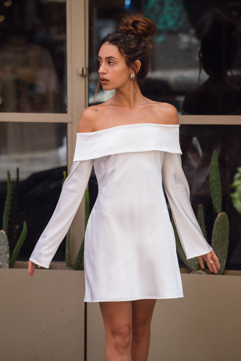 White satin off-shoulder mini dress with long sleeves, worn on-model near a storefront.