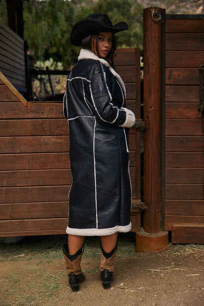 Black faux leather long coat with white sherpa trim, shown from behind at a ranch gate.