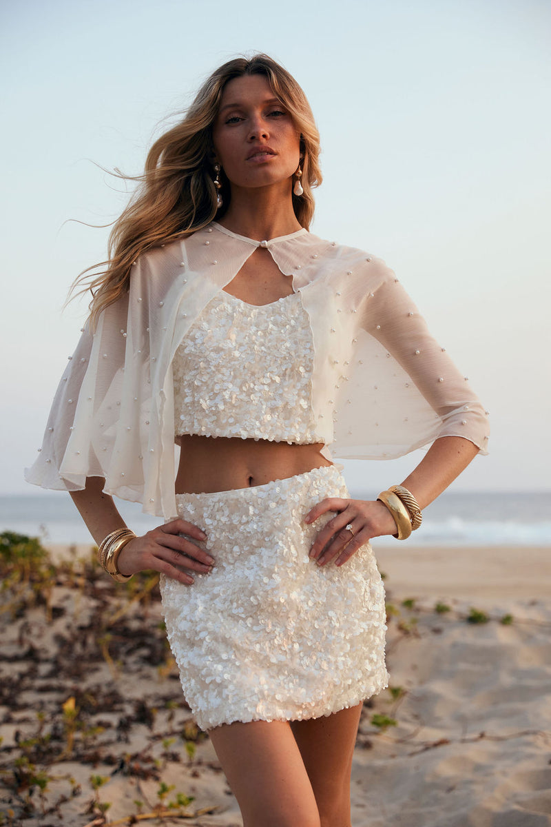 White sequin mini skirt styled with matching crop top and pearl cape on the beach.