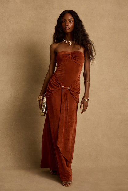 Model wearing the 12th Tribe Madonna Rust Maxi Dress, a sleek rust-toned stretch slinky maxi dress featuring a ruched bust, elegant front gold beaded and drape detail. Perfect for warm summer nights, vacation dinners, and nights out on the town.