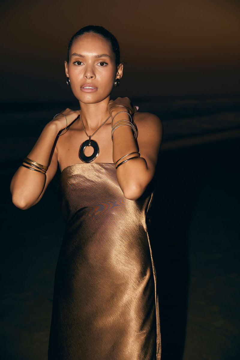 Bronze satin strapless maxi dress styled with gold cuffs and pendant at dusk.