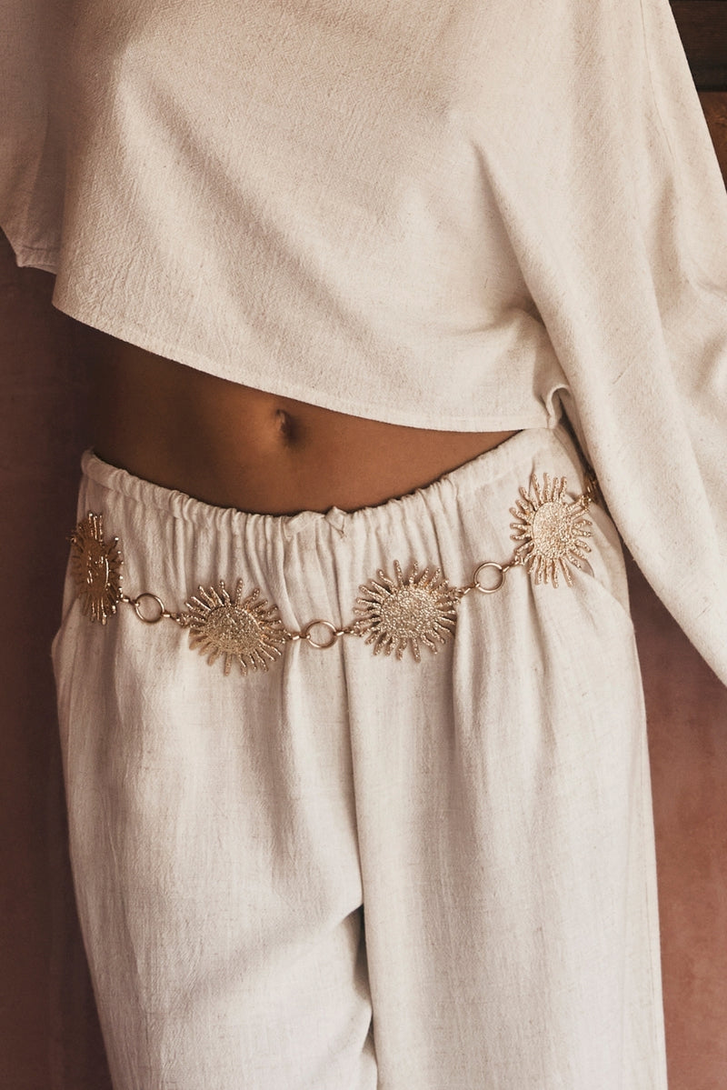 Gold sunburst chain belt worn over linen pants with a cream crop top.