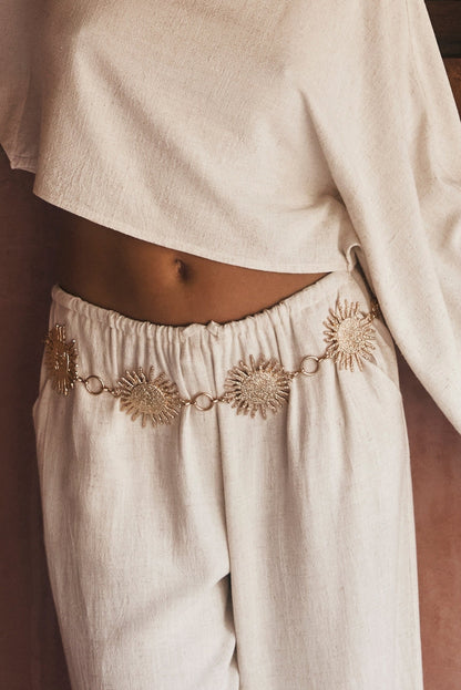 Gold sunburst chain belt worn over linen pants with a cream crop top.
